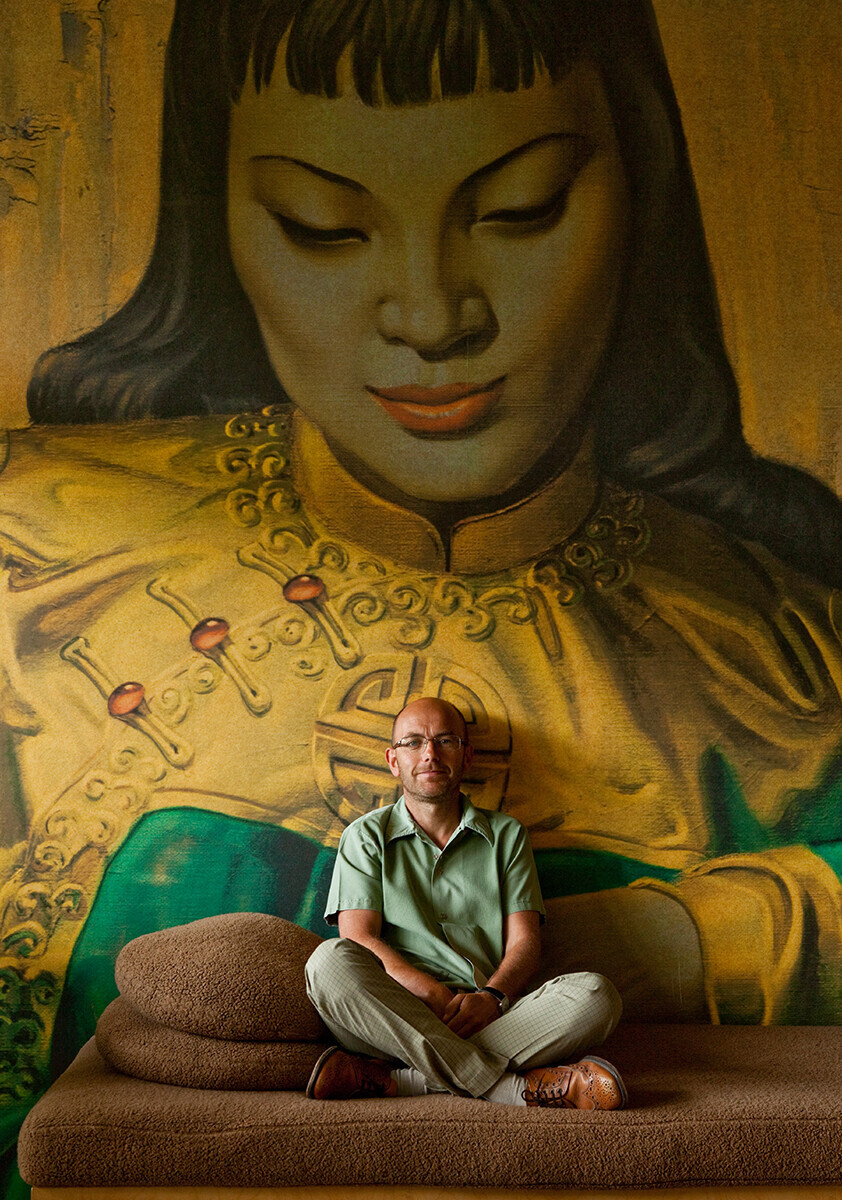 Wayne Hemingway sitting on a brown sofa in front of 'Lady Of The Orient' printed as a wallpaper mural