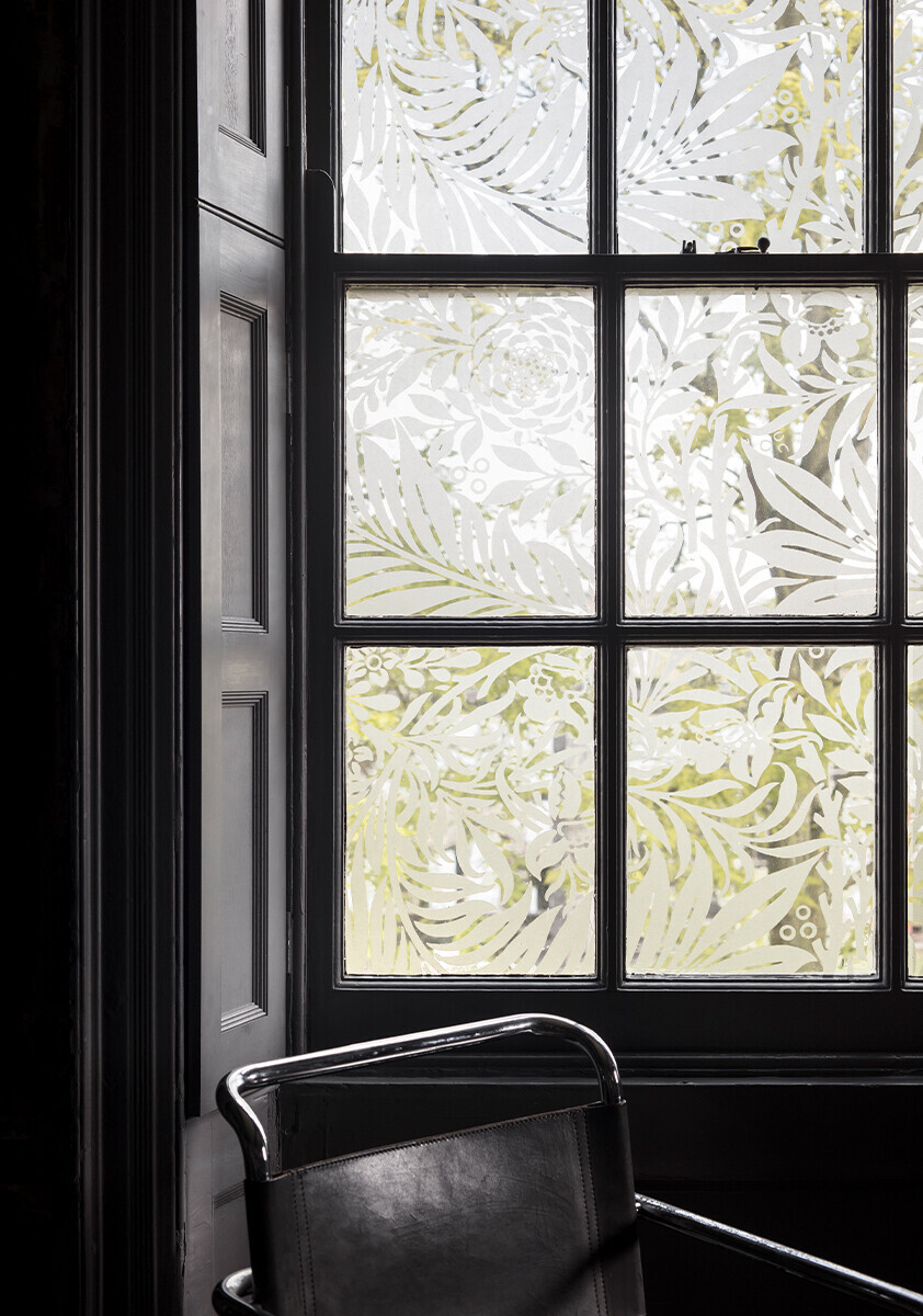 'Larkspur' by William Morris as a window film in a dark interior. 'Larkspur' depicts an ornate foliate design with flowers and leaves, in cool grey tones/